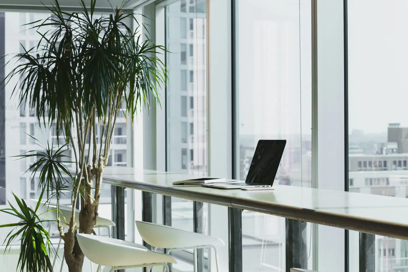 Espacio de trabajo luminoso con grandes ventanales, plantas y una laptop sobre una mesa alta