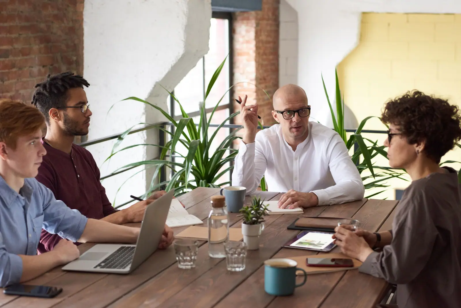 Equipo reunido alrededor de una mesa con laptops y plantas, conversando durante una reunión de trabajo